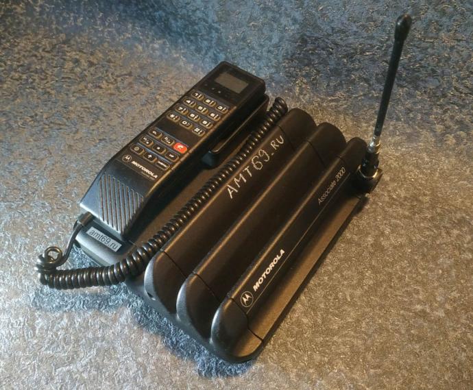 Винтажный сотовый телефон-чемодан Motorola Associate 2000 аналогового стандарта NMT-450 на темном рельефном фоне. Массивный черный пластиковый корпус с ребрами радиатора, проводной трубкой и штыревой антенной.